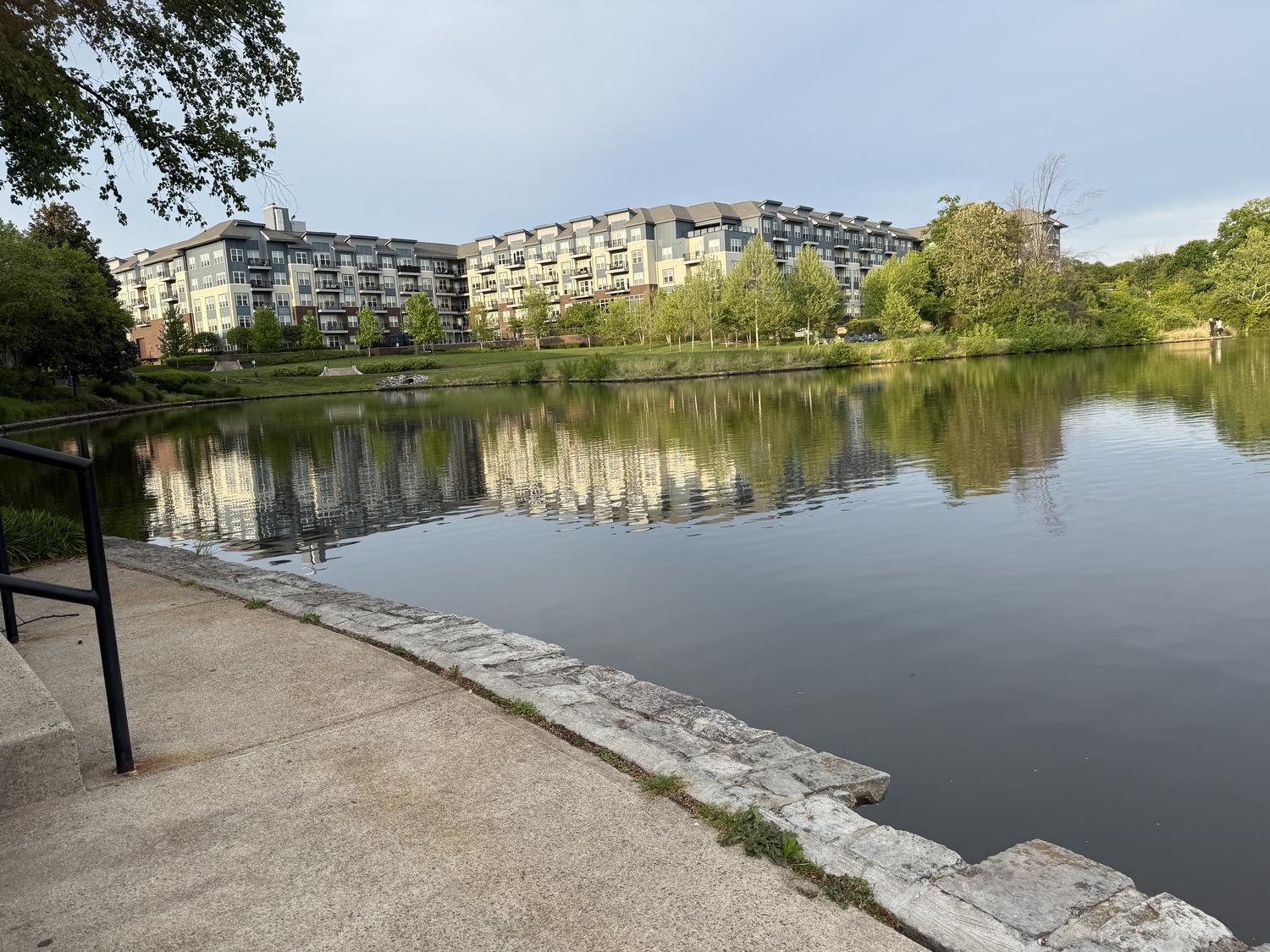 A concrete path and handrail leading down to a cut-stone pond edge, with apartment buildings reflected in the water beyond.